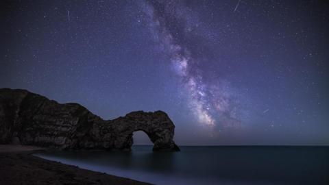 The Milky Way can be seen in the night sky over the Dorset Coast. It is seen as a streak of white across the dark sky full of stars. The sea is a dark blue. In the foreground is a dark beach in front of the natural arch of Durdle Door