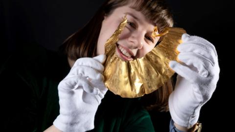 A person is holding a delicate, golden, fan-shaped object called a lunula with both hands. They are wearing clean white gloves. The object shines under the light, with a textured surface. The person holding the lunula is dressed in a dark green top and wearing a blue wristwatch on their left wrist. The background is dark, which makes the golden object and the gloved hands stand out clearly.