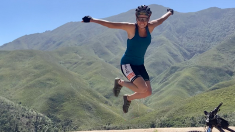 Zelda Wilson jumping above a wall on a cycling challenge. She is smiling and has her arms in the air. Behind her are large green hills