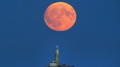 A red-orange full Moon hangs low in the night sky over a lighthouse