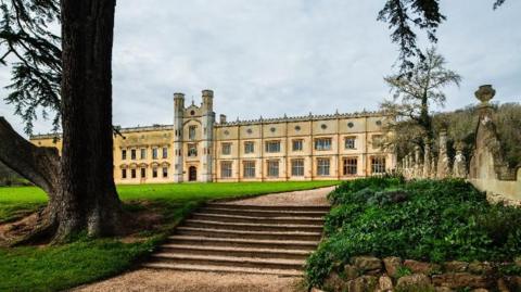 Ashton Court Mansion, a three-storey yellow 14th building with two towers at each side of the entrance is surrounded by trees and bushes.