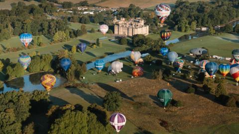 More than 20 hot air balloons of different colours and sizes ascending above Longleat Estate, with the Longleat house below and a body of water running through the centre of the fields.