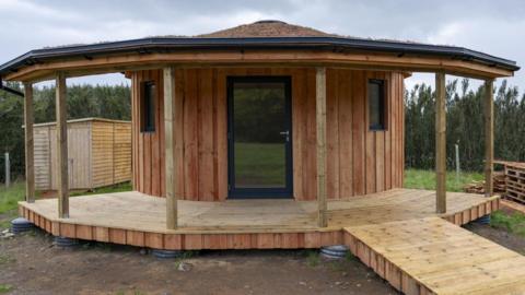 The centre has a round-shaped wooden building with a glass front door and windows in the walls. Around the building is a wooden, veranda-style structure. Trees surround the site.