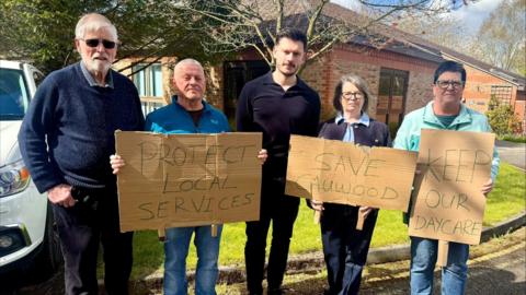 Five people stand outside a brick building holding handmade cardboard signs that read “Protect Local Services,” “Save Cauwood,” and “Keep Our Daycare,” taking part in a small community protest.