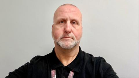 A man with very short hair and white stubble is standing in front of a blank wall, wearing a black shirt.