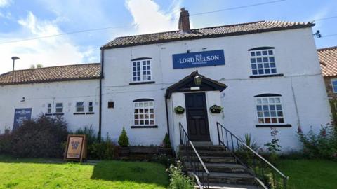 A large white building is pictured, with four windows and a protruding porch with a its own small triangular roof. There's hanging baskets on either side of the porch door, a light on the roof, and a blue sign saying 'The Lord Nelson Poppleton' above it. To the left there's an extension of the building, with four smaller windows and another blue The Lord Nelson Poppleton sign. There's stairs leading up to the door with hand railings on either side, and a green lawn at the front.