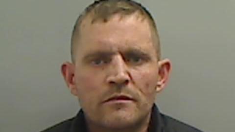 Mugshot of Dobie. He is clean shaven with short fair hair and has a menacing expression on his face.
