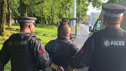 Two police officers in black uniform hold the arms of a man wearing handcuffs as they guide him towards a police van parked at a roadside.