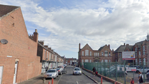 Worcester Street lined with red brick terrace houses and cars to the left. A large red brick primary school can be seen to the right.