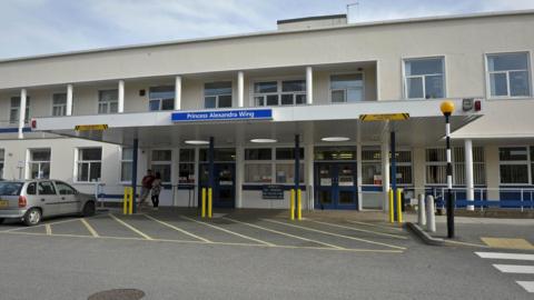 Royal Cornwall Hospital frontage with PRINCESS ALEXANDRA WING written above the main door on a blue sign. There are two people stood outside the two storey white building speaking. 