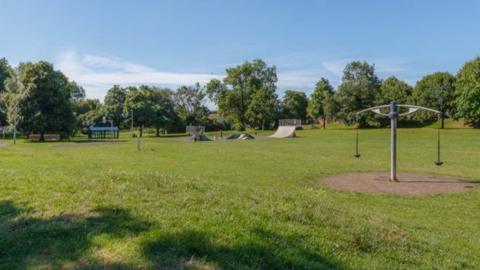A park with a large grass area with a skate ramp, benches, shelter and a piece of play equipment within the grass. It is a sunny day with a blue sky and there are trees in the distance around the edge of the grass