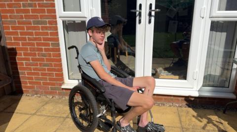 A young man in a blue cap and shorts and a grey tshirt sitting in a wheelchair, in front of some patio doors, with his hand supporting his head 