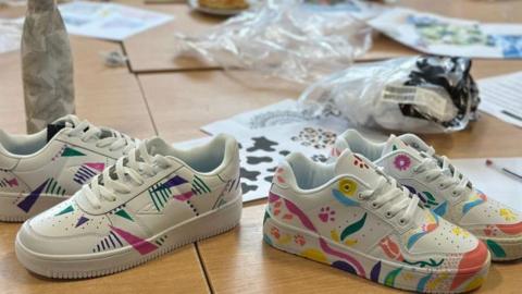 Two pairs of white trainers perched on a table, they have been decorated with colourful drawings including pink, purple and green triangles and flowers and leaves