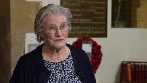 Alison Hoddell stands in the church with the plaque and a poppy wreath behind her. She has short grey hair and is wearing a blue patterned dress and navy cardigan. 