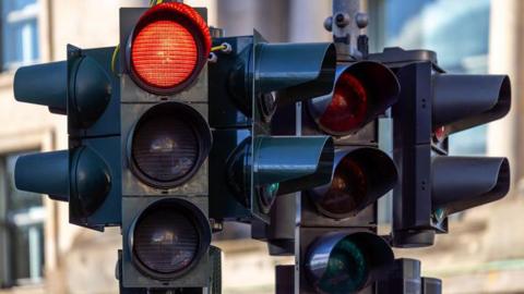 A close-up of several traffic lights mounted on a metal pole at a street junction. The primary traffic light in the foreground is clearly visible, and its top circular light is illuminated bright red. The red light is housed within a black rectangular casing, with two unlit circular lights positioned below it for the amber and green signals.