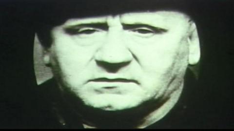 A close-up, black-and-white photograph of Garvin. He is facing directly toward the camera and is wearing a dark hat that sits low on the head.
The background is very dark and lacks any visible detail, which draws attention to the face. The image has a grainy texture and high contrast.