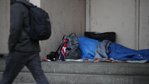 A person is pictured in a blue sleeping bag with bags surrounding their head. They are sleeping on the steps to a building as a man walks past.