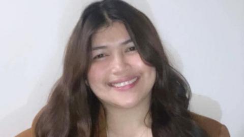 A close-up image of Cathrene Kith Quiñones Singco. She is a young woman, with long, dark wavy hair and she is smiling at the camera. She is wearing a brown jacket and is standing in front of a white wall.