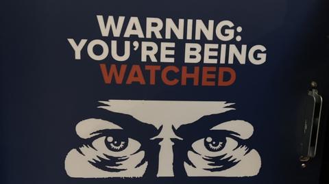 Sign with the words "warning you're being watched" above a drawing of eyes