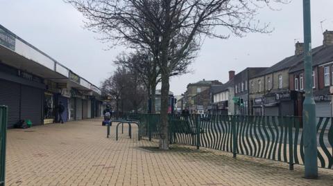 An empty high street. Many of the shops have shutters down. There are no people walking around.