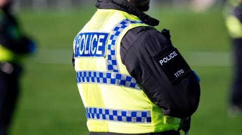 A generic shot of a police officer stood with his back turned. His head cannot be seen. He is wearing a hi-viz vest with 'Police' on the back. 
