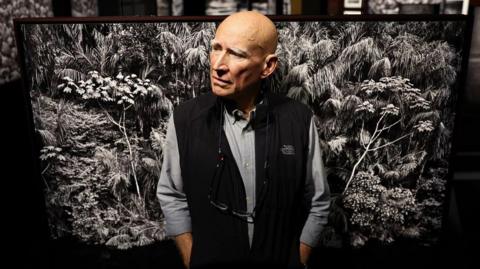 Sebastião Salgado standing in front of a black and white photo of trees in the Amazon