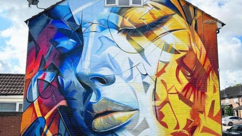 Colourful mural of a woman's face with closed eyes. Colours range from red and purple on the left to blue to yellow and orange on the right. The woman's face is blue, starting out more saturated on the left and becoming more fainted towards the right with her lips and left eye coloured yellow.