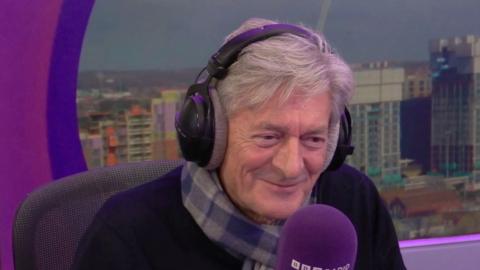 An older man with a checked scarf and grey hair sits in a radio studio with headphones on