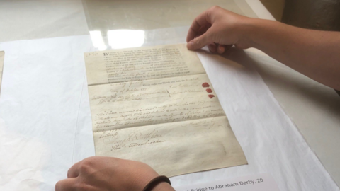 A pair of hands holding the corners of a piece of paper with some writing on it, on a white table top