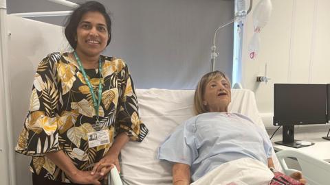 Senior care worker Sajee Gunarathna stands next to the bedside of a model patient called 'Dorris'. Sajee is wearing a black and yellow floral top and has long black hair. 