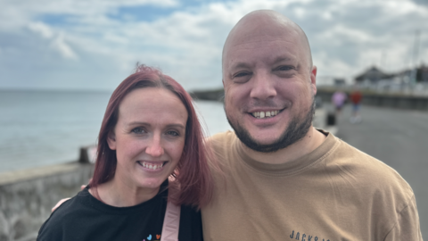 Kayleigh Avery, with red dyed hair and wearing a black t-shirt, stands next to her husband David, who is wearing a brown t-shirt.