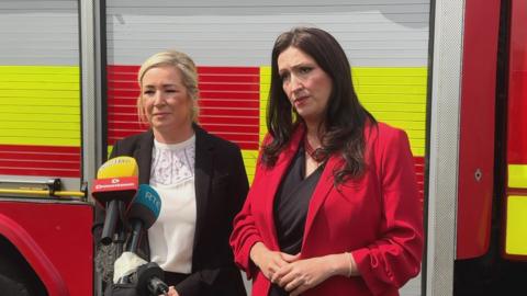 Michelle O'Neill and Emma Little-Pengelly addressing reporters at the official opening of the Northern Ireland Fire and Rescue Service Learning and Development College near Cookstown. O'Neill has shoulder-length blonde hair, tied back and is wearing a black suit jacket over a white top. Little-Pengelly has long, dark hair and is wearing a red suit jacket over a black top or dress. They are standing in front of a fire engine.