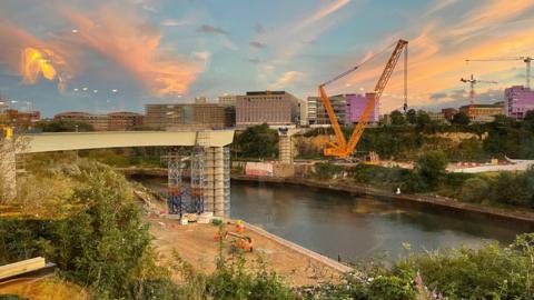 A photo at sunset showing the construction of a bridge, with the structure stopping just before the river. It is covered in scaffolding and a crane can be seen on the other side. 