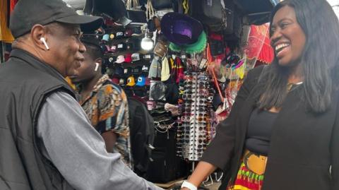 A woman in a black jacket and top and a colourful yellow skirt smiles as she shakes hands with an older man. They are standing in an outdoor market and there are rows on sunglasses and hats for sale. 