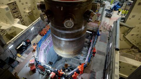 A huge steel cylinder is lowered into a big concrete cavity beneath it. Workers in orange high vis watch on, in a large building made of cream concrete