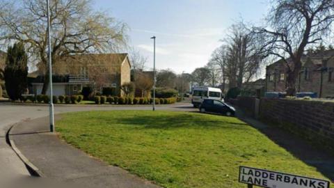 A green patch of grass on the side of a housing estate. On the bottom right is a road sign that reads: Ladderbanks Lane