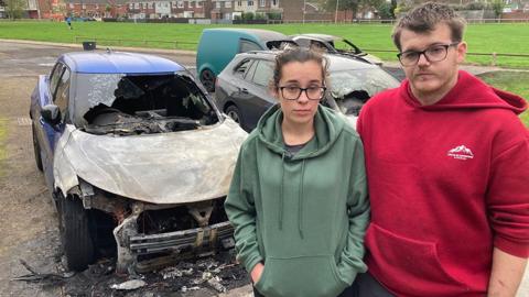 Shannon Rex and her partner Luke Deekens stand in front of their two burnt out cars. She has long dark hair pinned back behind her head and wears glasses and a green hooded top. He has short dark hair and wears glasses and a red hooded top.