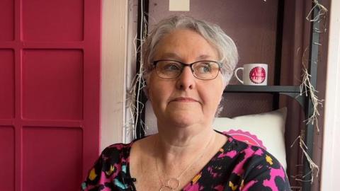 Lindsey Brown, who has short grey hair, sitting in front of a black, metal book case with fairy lights wound round it. She's wearing black glasses,  a top with a bright neon splatter pattern on it and a silver necklace.