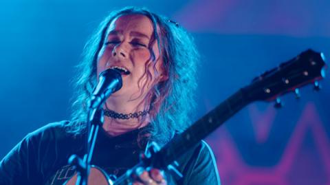 Nati has her eyes closed as she sings into a microphone and plays a guitar. She has curly hair down to her shoulders. The stage inside the festival's big top is blue and pink.