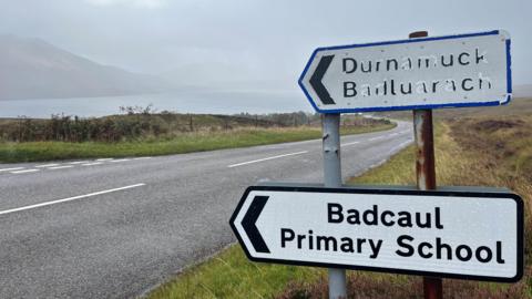 A sign pointing towards Badcaul Primary School. It has black writing on a white background and is in front of a road on a grass verge.