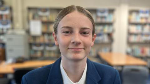 A teenage girl, with brown hair slicked back in a middle parting and tied back. She has a navy jacket and a white tshirt