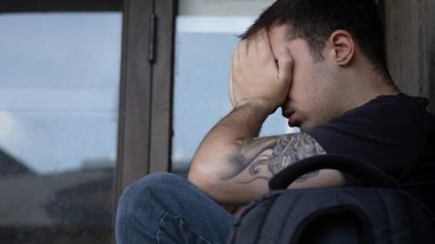 a young man holding his head in his hands wearing a black t-shirt. He is sitting in front of a glass door