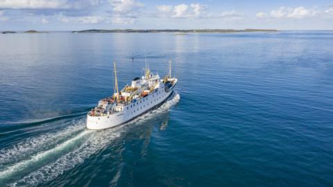 Scillonian III sails between Penzance and the Isles of Scilly on a sunny day. One of the islands is in the distance. Large groups of passengers can be seen on the vessel's decks.