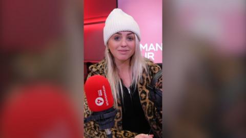 Aimee Fuller is wearing a white bobble hat and a leopard print jacket over a black top.