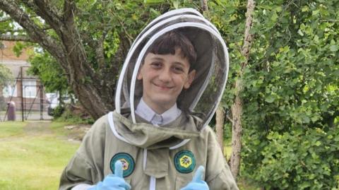 A boy wearing a beekeeping suit. He is stood next to a bush in a field.