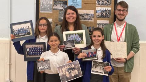 Five people are standing in front of a board displaying old photographs on a wall. Ffion, a pupil has brown hair and black glasses, she is holding two colour photographs of school children. Caia has brown hair tied back and is holding two black and white photos of schoolchildren. Emilee has long brown hair and is holding one black and white school photos and one in colour. Headteacher Hannah Gharu has brown hair and is holding a black and white school photo and a colour school photo. Tom Ollivier has brown hair and glasses, with a green cardigan. He is holding an old log book