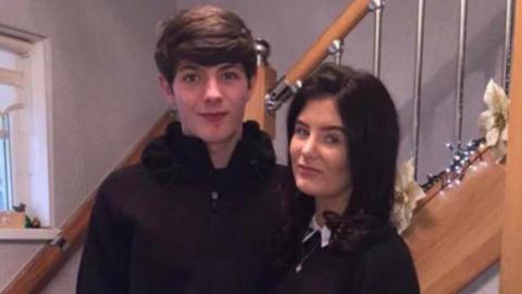 Kasey Anderson has short brown hair and is wearing a black hoodie. He is stood with Molly Anderson who had black hair and is wearing a black top with a white shirt and silver pendant necklace. 