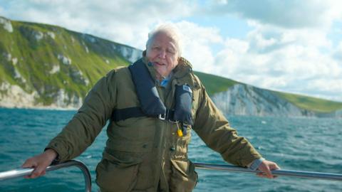 Sir David Attenborough, an older man with white hair, is pictured at the bow of a boat on the water. He is wearing a large green coat and black life jacket and is gripping a metal railing with each of his hands. Behind him, large white cliffs rise out of the water. 