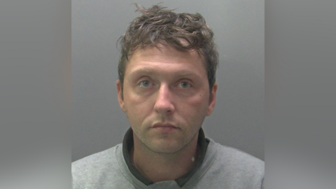 Custody photo of Craig France against a grey background. He is wearing a light grey sweatshirt while looking at the camera with a straight face. He has brown curly hair.

