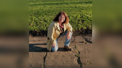 Dr Myra Giesen, a woman with shoulder length brown hair worn down and glasses, kneels on a flat, inscribed gravestone as she smiles at the camera